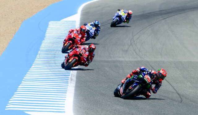 Racing motorcycles lean into a curve on a race track, multiple riders in colorful gear leading the pack.
