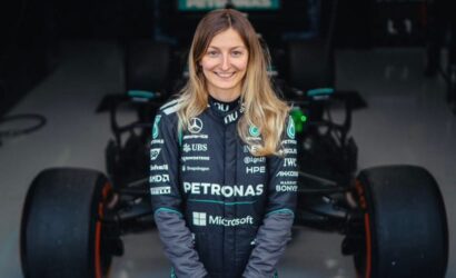 Female Formula 1 driver in Petronas team suit standing in a garage with a race car behind her.