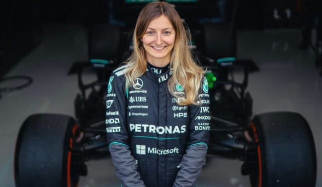 Female Formula 1 driver in Petronas team suit standing in a garage with a race car behind her.