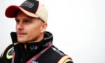 Portrait of a male race car driver wearing a beige cap with a red brim and a black jacket covered in sponsor logos, looking off to the left.