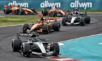 A pack of Formula 1 race cars cornering on a track, with Allwyn banners visible in the background.