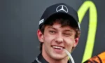 Young man wearing a black Mercedes cap, smiling with a strap in his mouth, in a racing environment background.