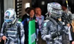 Two Mercedes Formula One drivers in dark racing suits and helmets walk through a garage, one carrying a handheld camera, with team members in the background.