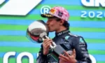 Race car driver in a dark blue suit and pink cap kissing a silver trophy on a podium after a victory.