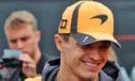 Smiling man wearing an orange cap with a black logo, with a photographer in the blurred background.