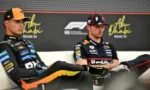 Two motorcycle racers sit on a stage at a press conference, wearing team sponsorship suits; one wears an orange-black cap, the other a dark cap, with sponsor banners behind them.