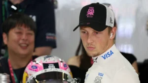 Male race car driver in a white racing suit with Visa branding, wearing a black cap with a pink emblem, looking to the right, with a pink-design helmet in front of him.