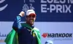 Male race winner on stage, wearing cap and sunglasses with a green cape, celebrating at a Formula E event (Hankook sponsor).