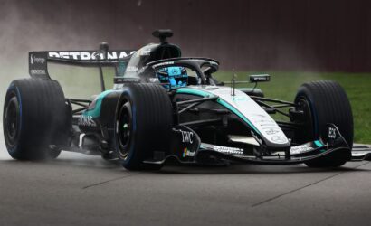 Mercedes-AMG Petronas Formula 1 car on a wet track, spraying water as it cornering at speed; teal-black livery and driver in blue helmet visible.