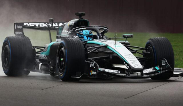 Mercedes-AMG Petronas Formula 1 car on a wet track, spraying water as it cornering at speed; teal-black livery and driver in blue helmet visible.