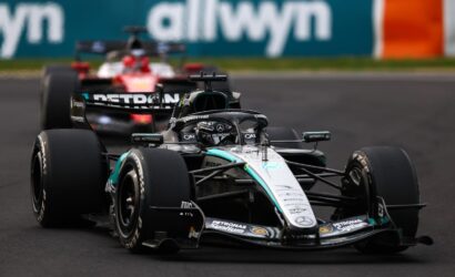 Mercedes F1 car leading on a race track, with a second car following and a blurred sponsor barrier in the background