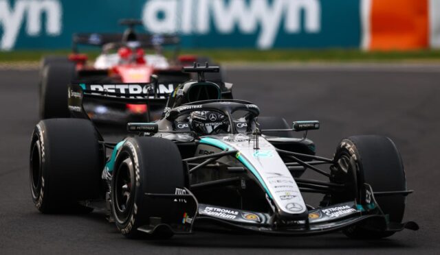 Mercedes F1 car leading on a race track, with a second car following and a blurred sponsor barrier in the background