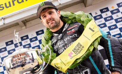 Male motorcycle racer wearing a laurel wreath and cap, holding a large silver trophy on a podium backdrop with sponsor banners.