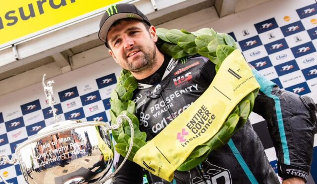 Male motorcycle racer wearing a laurel wreath and cap, holding a large silver trophy on a podium backdrop with sponsor banners.
