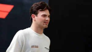 Man with dark hair wearing a white crewneck sweater stands outdoors, looking to the right with a neutral expression against a dark background with a red shape nearby.