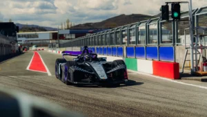 Black race car with purple accents on a track, near pit barriers and a green signal light under a cloudy sky.