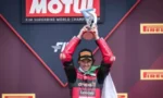 Race car driver on a podium raising a trophy in victory, wearing a red racing suit with sponsor logos, with Motul and Pirelli banners in the background.