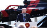 Speaker in a suit delivers a speech at a podium with the Turkish emblem, a red-black helicopter in the background behind him.