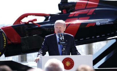 Speaker in a suit delivers a speech at a podium with the Turkish emblem, a red-black helicopter in the background behind him.