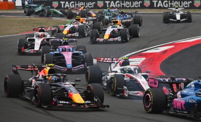 A dense pack of Formula 1 cars navigating a left-hand turn on a race track, colorful sponsor liveries visible with TAG Heuer banners in the background.