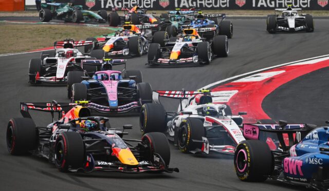 A dense pack of Formula 1 cars navigating a left-hand turn on a race track, colorful sponsor liveries visible with TAG Heuer banners in the background.