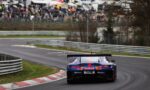 Blue and red Red Bull race car seen from the rear speeding through a left-hand curve on a race track, with a crowd watching behind barriers in the background.