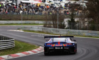 Blue and red Red Bull race car seen from the rear speeding through a left-hand curve on a race track, with a crowd watching behind barriers in the background.