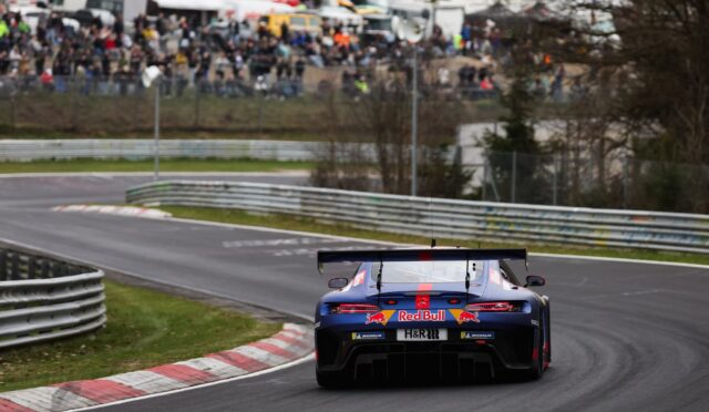 Blue and red Red Bull race car seen from the rear speeding through a left-hand curve on a race track, with a crowd watching behind barriers in the background.
