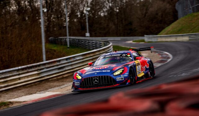 Red Bull–wrapped Mercedes-AMG GT3 racing car rounding a track curve, headlights on, barrier wall to the left.