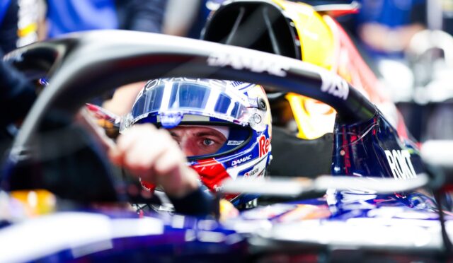 Close-up of a Formula 1 driver wearing a blue helmet inside a race car cockpit, sponsors visible on suit and car.