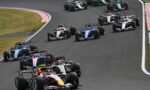 Several Formula 1 cars race around a left-hand corner in a tight formation on a asphalt track.