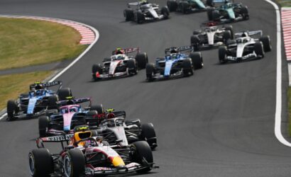 Several Formula 1 cars race around a left-hand corner in a tight formation on a asphalt track.