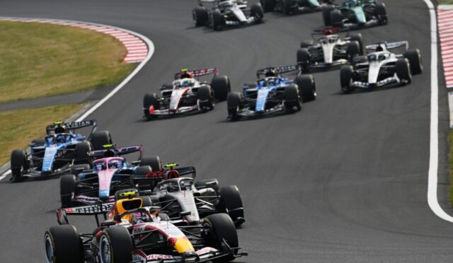 Several Formula 1 cars race around a left-hand corner in a tight formation on a asphalt track.