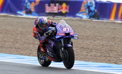 Professional motorcycle racer leaning into a turn on a purple bike with Red Bull helmet, sponsors visible on suit and bike, track barriers in background.