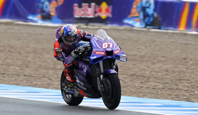 Professional motorcycle racer leaning into a turn on a purple bike with Red Bull helmet, sponsors visible on suit and bike, track barriers in background.