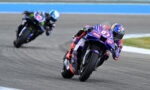 MotoGP rider leaning into a turn on a race track; purple bike with sponsor logos.