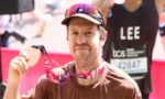 Male runner displaying his finisher medal after a race, wearing a brown shirt and cap, with bibs and spectators in the background.