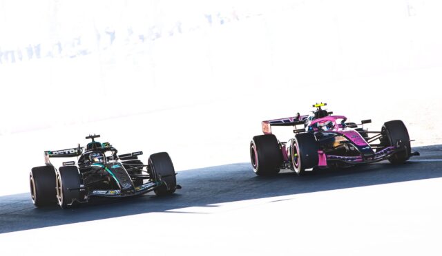 Two Formula 1 cars race side by side on a sunlit track, a dark Mercedes on the left and a pink Racing Point on the right.