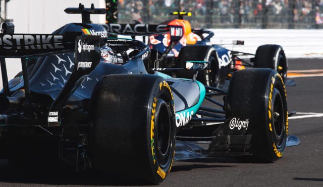 Two Formula 1 cars race side by side on a track, rear tires smoking as they squeeze past each other with a blurred crowd in the background.