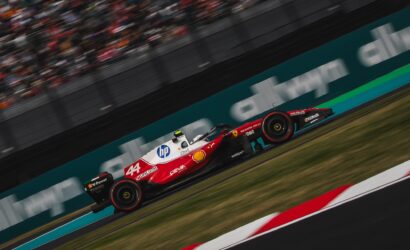Red Formula 1 race car (number 44) speeding along a race track with sponsor logos visible (Unicredit, HP, Shell) and blurred grandstands in the background, motion suggesting high speed.