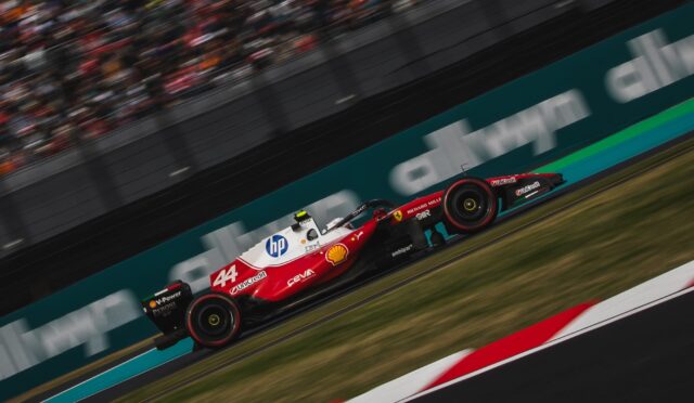 Red Formula 1 race car (number 44) speeding along a race track with sponsor logos visible (Unicredit, HP, Shell) and blurred grandstands in the background, motion suggesting high speed.