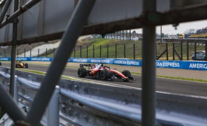 Red Formula 1 car racing on track, seen through metal barriers with spectators in the stands in the distance.