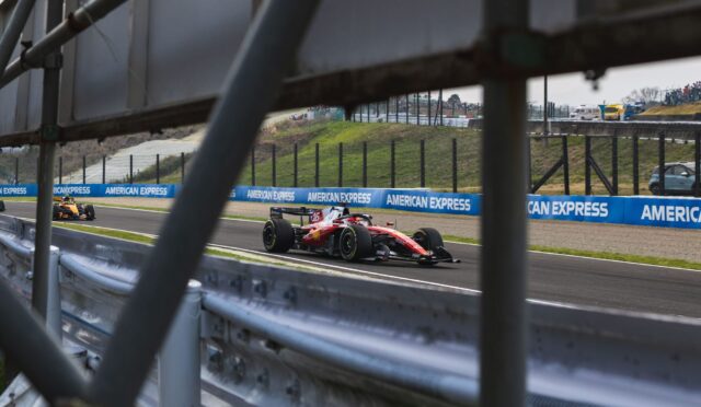 Red Formula 1 car racing on track, seen through metal barriers with spectators in the stands in the distance.