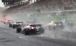 Formula One cars racing off the start in heavy smoke, crowded grandstands in background, wet track conditions.