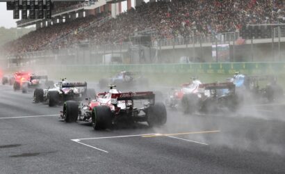 Formula One cars racing off the start in heavy smoke, crowded grandstands in background, wet track conditions.