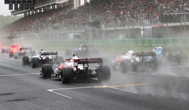 Formula One cars racing off the start in heavy smoke, crowded grandstands in background, wet track conditions.