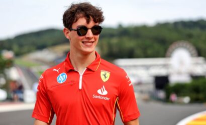 Young man in a red Ferrari polo with sponsor logos and sunglasses at a racetrack, smiling.