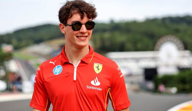 Young man in a red Ferrari polo with sponsor logos and sunglasses at a racetrack, smiling.