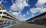 Wide view of a race track flanked by white pit buildings, sponsor banners reading 'crypto.com' along the barriers, under a bright blue sky with white clouds.