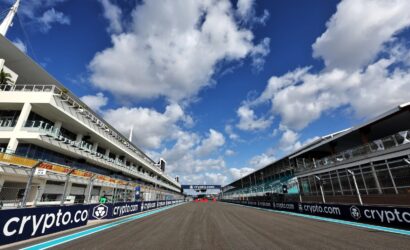 Wide view of a race track flanked by white pit buildings, sponsor banners reading 'crypto.com' along the barriers, under a bright blue sky with white clouds.
